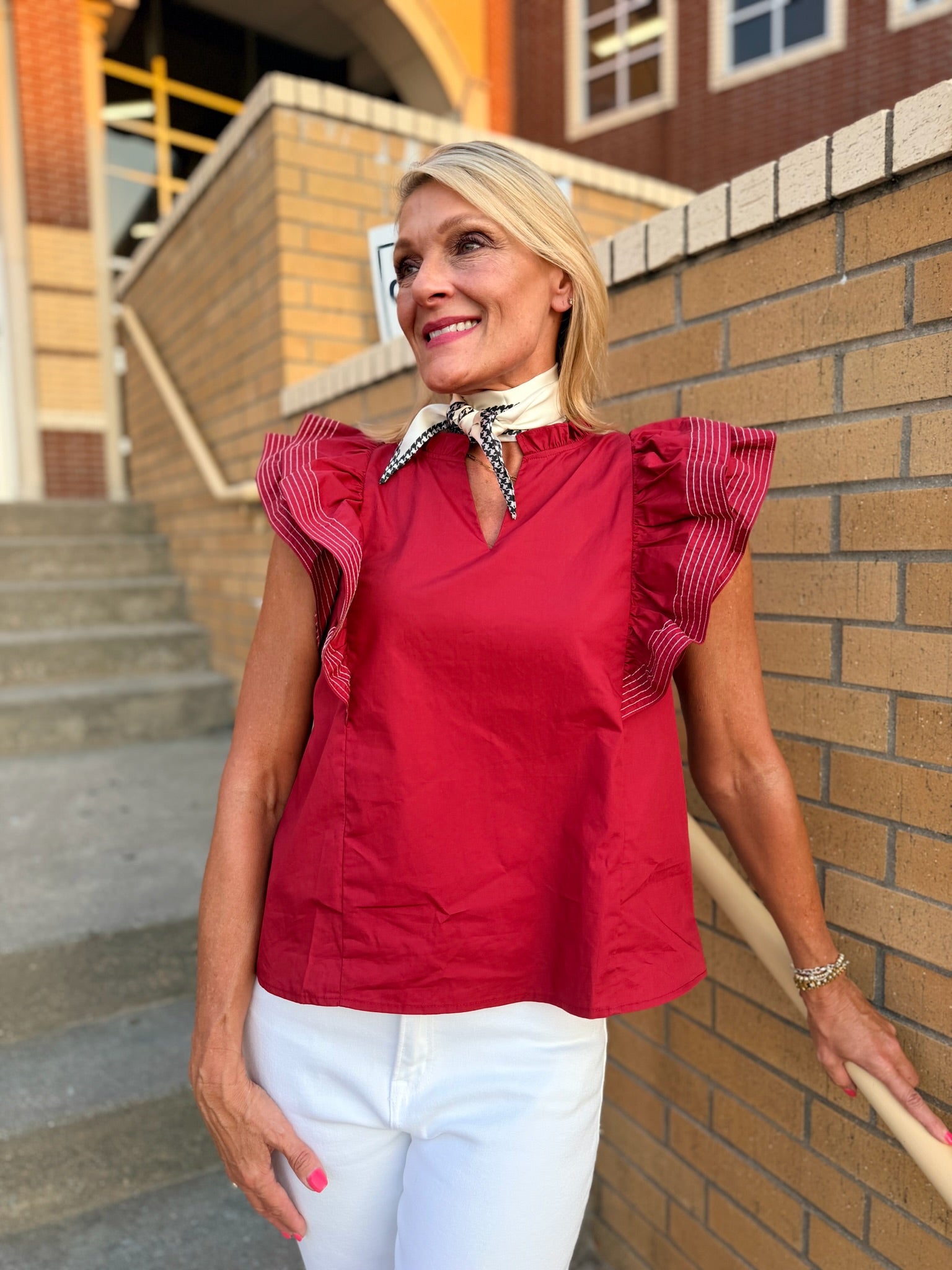 A woman wearing a garnet ruffled sleeveless blouse with a cream and houndstooth scarf detail and white pants, standing outdoors against a brick wall and staircase.