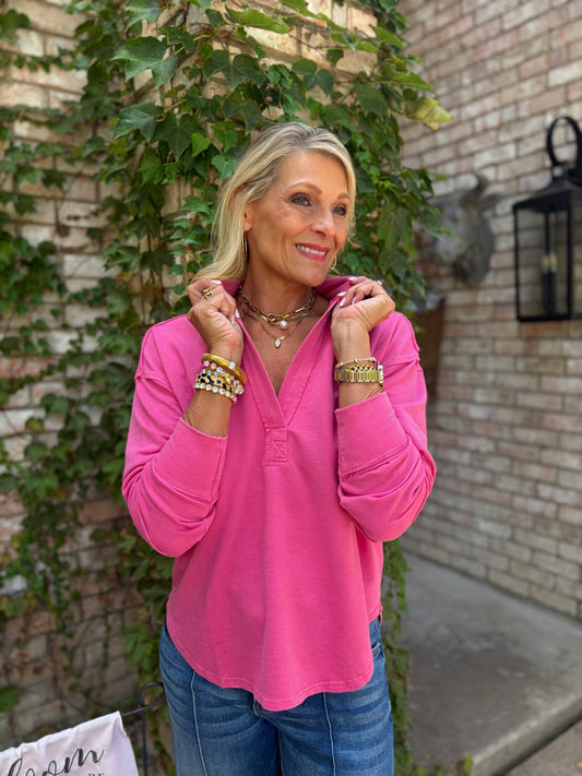 A woman wearing a bright pink long-sleeve henley top and blue jeans, standing outdoors in front of a brick wall with green ivy. She is smiling and adjusting the collar of the top, accessorized with layered necklaces and bracelets.