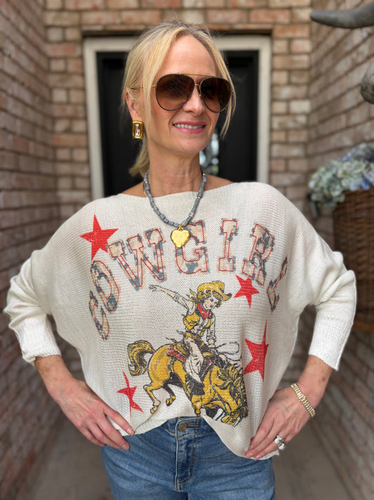 A woman wearing a cream-colored knit sweater with a cowgirl graphic and the word 'COWGIRL' printed across the front, paired with blue jeans, standing in front of a brick wall.