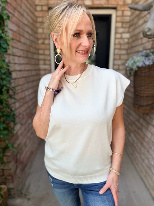 A woman wearing a white-colored short-sleeve knit top and blue jeans, standing in a brick alleyway.