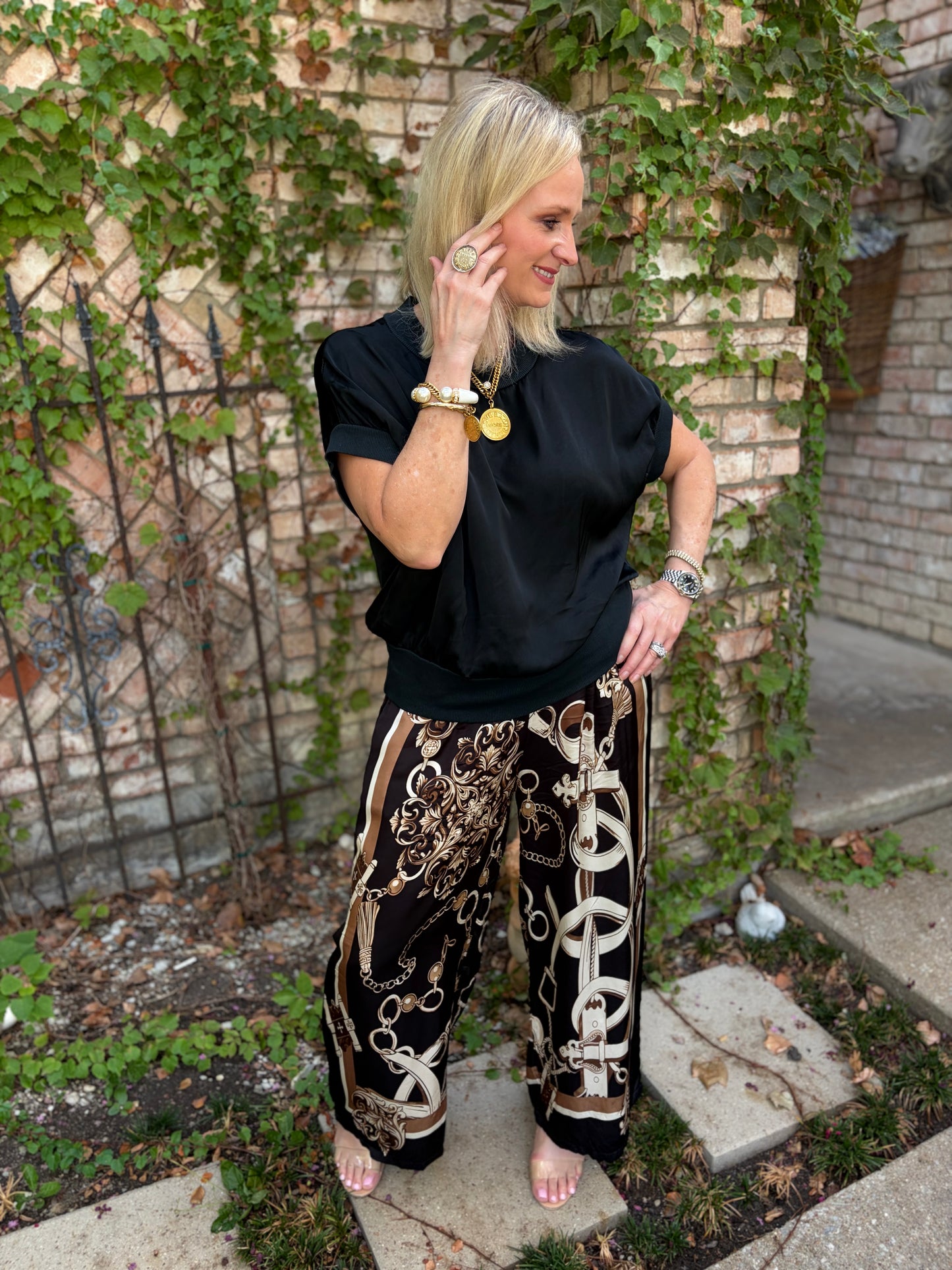 A woman stands outdoors in front of a brick wall covered in green ivy, wearing a black short-sleeve top and wide-leg black pants with a beige and gold chain-link print. She is barefoot and accessorized with gold jewelry.