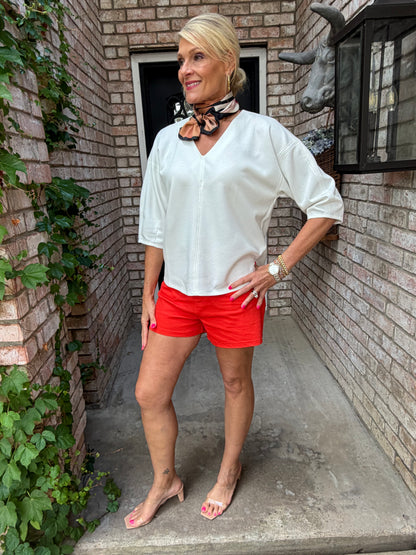 Woman in a white blouse and red shorts standing in front of a brick wall with greenery.