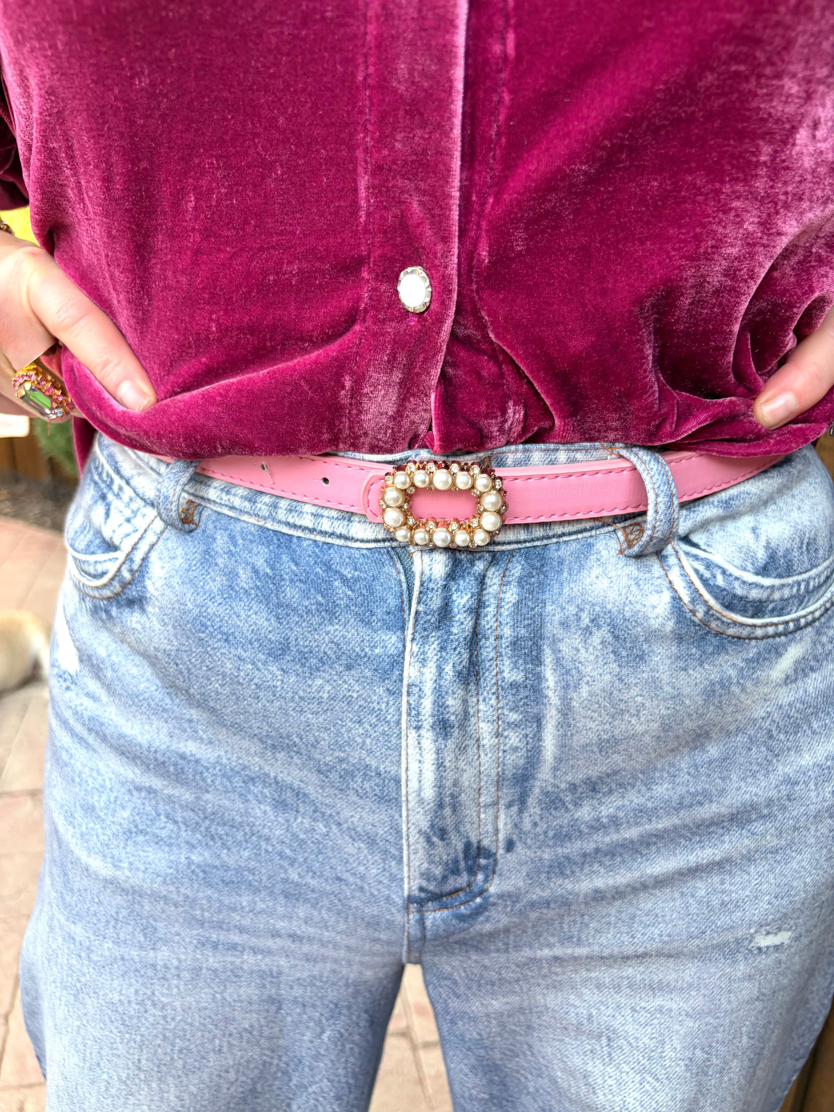 a light pink skinny leather belt with a decorative rhinestone and pearl buckle, paired with light blue jeans and a magenta velvet top.