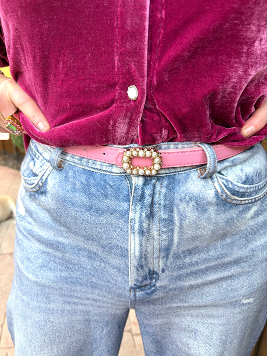  a light pink skinny leather belt with a decorative rhinestone and pearl buckle, paired with light blue jeans and a magenta velvet top.