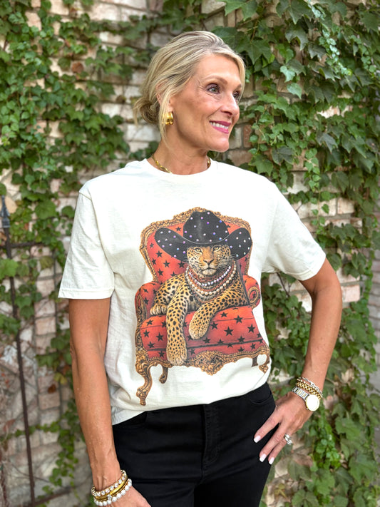 A woman wearing a cream-colored graphic t-shirt with a leopard sitting on an orange armchair wearing a black, blinged cowboy hat design, paired with black pants, standing in front of a brick wall covered in green ivy.