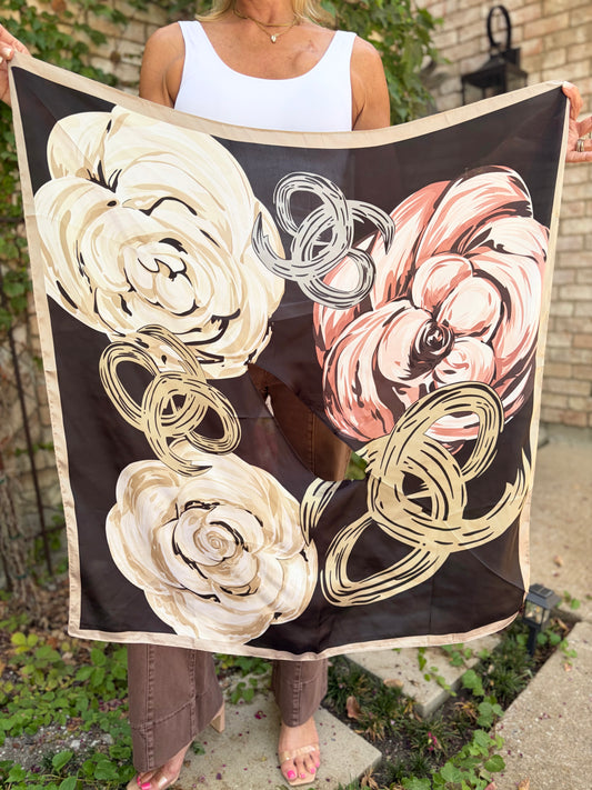 A person outdoors holds up a square scarf with a black background and large floral print in cream, blush pink, and beige