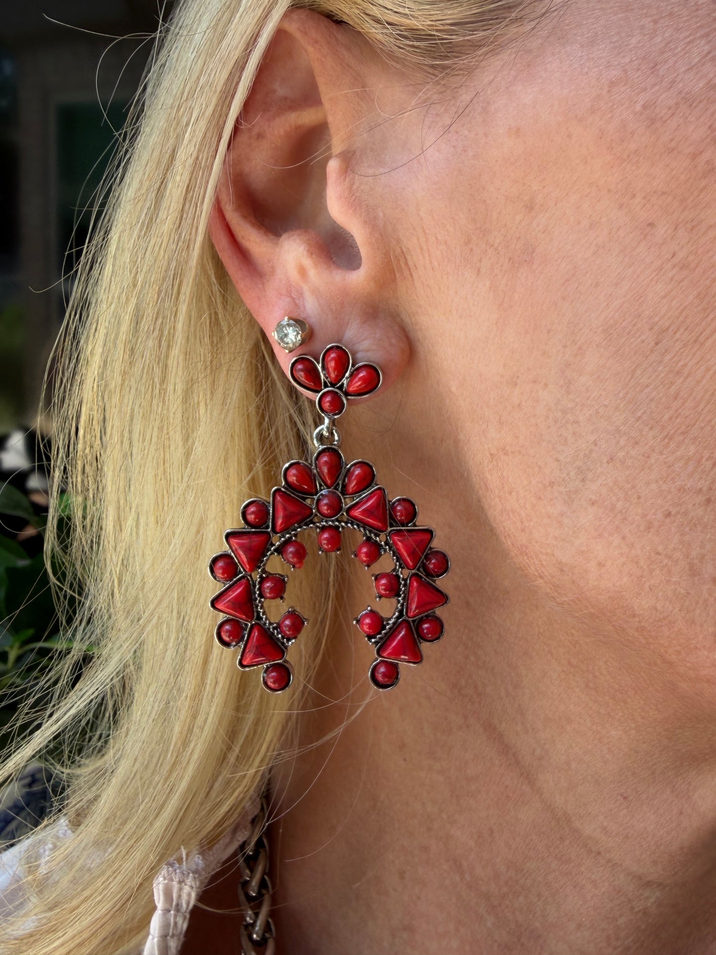 A close-up of a person's ear wearing a large, intricate red and dark silver beaded western style earring with a small silver stud earring underneath, with blonde hair framing the side of the face.