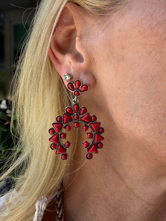 A close-up of a person's ear wearing a large, intricate red and dark silver beaded western style earring with a small silver stud earring underneath, with blonde hair framing the side of the face.