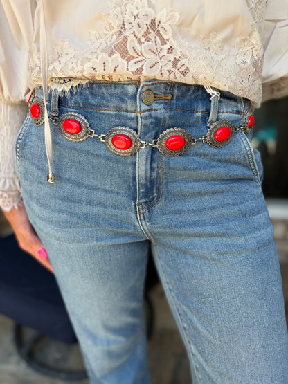 Close-up of a person wearing light blue jeans with a decorative chain belt featuring large red oval stones and silver-toned metal accents, paired with a cream lace blouse.