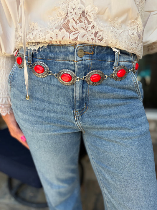 Close-up of a person wearing light blue jeans with a decorative chain belt featuring large red oval stones and silver-toned metal accents, paired with a cream lace blouse.