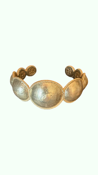 An antique-style gold cuff bracelet featuring five large, textured oval panels connected by beaded borders, set against a solid off-white background.