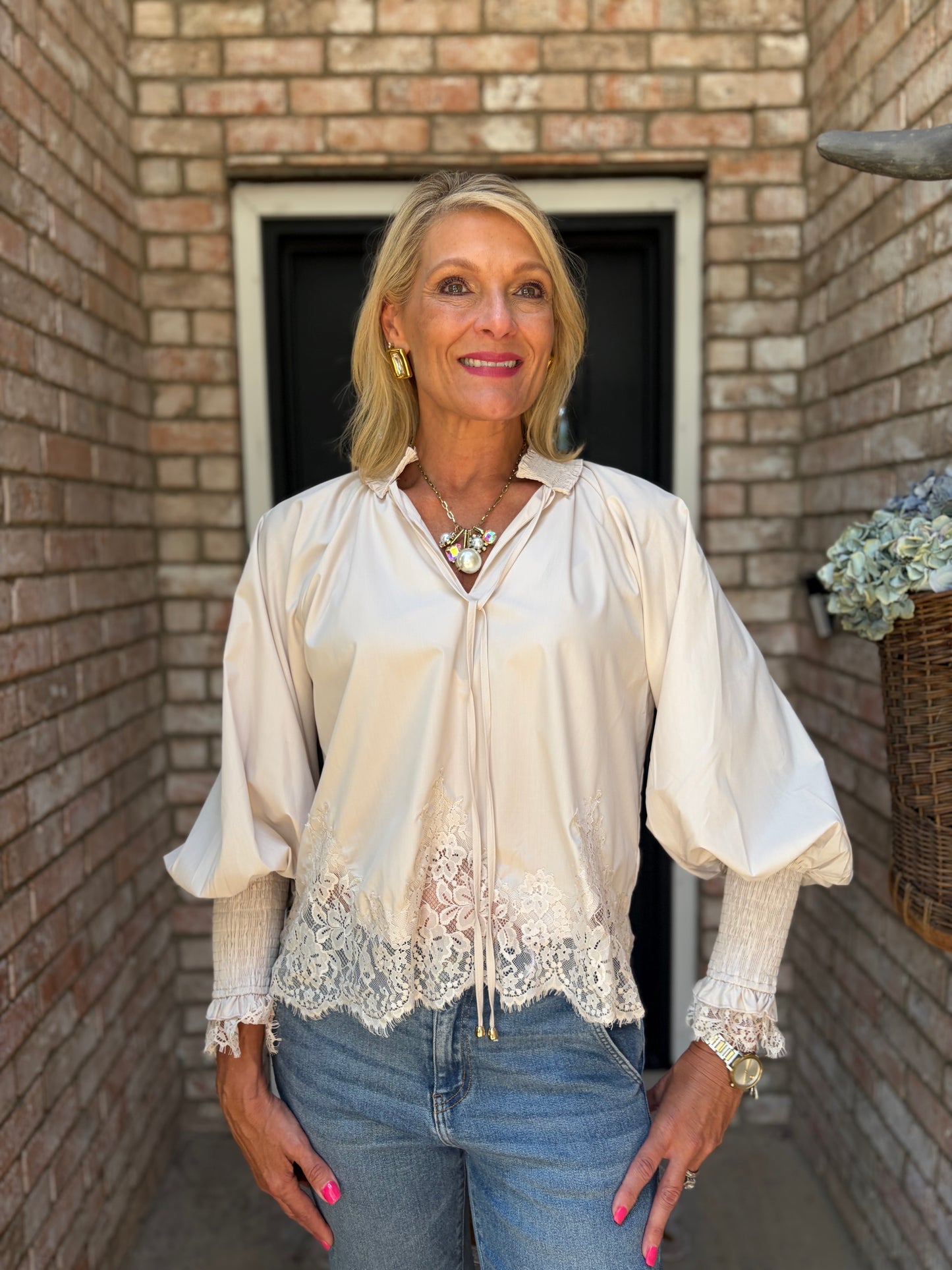 A woman wearing a pearl-colored satin blouse with lace trim and puff sleeves, paired with blue jeans, standing in front of a brick wall doorway.