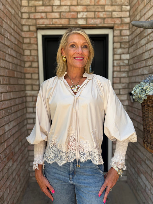 A woman wearing a pearl-colored satin blouse with lace trim and puff sleeves, paired with blue jeans, standing in front of a brick wall doorway.