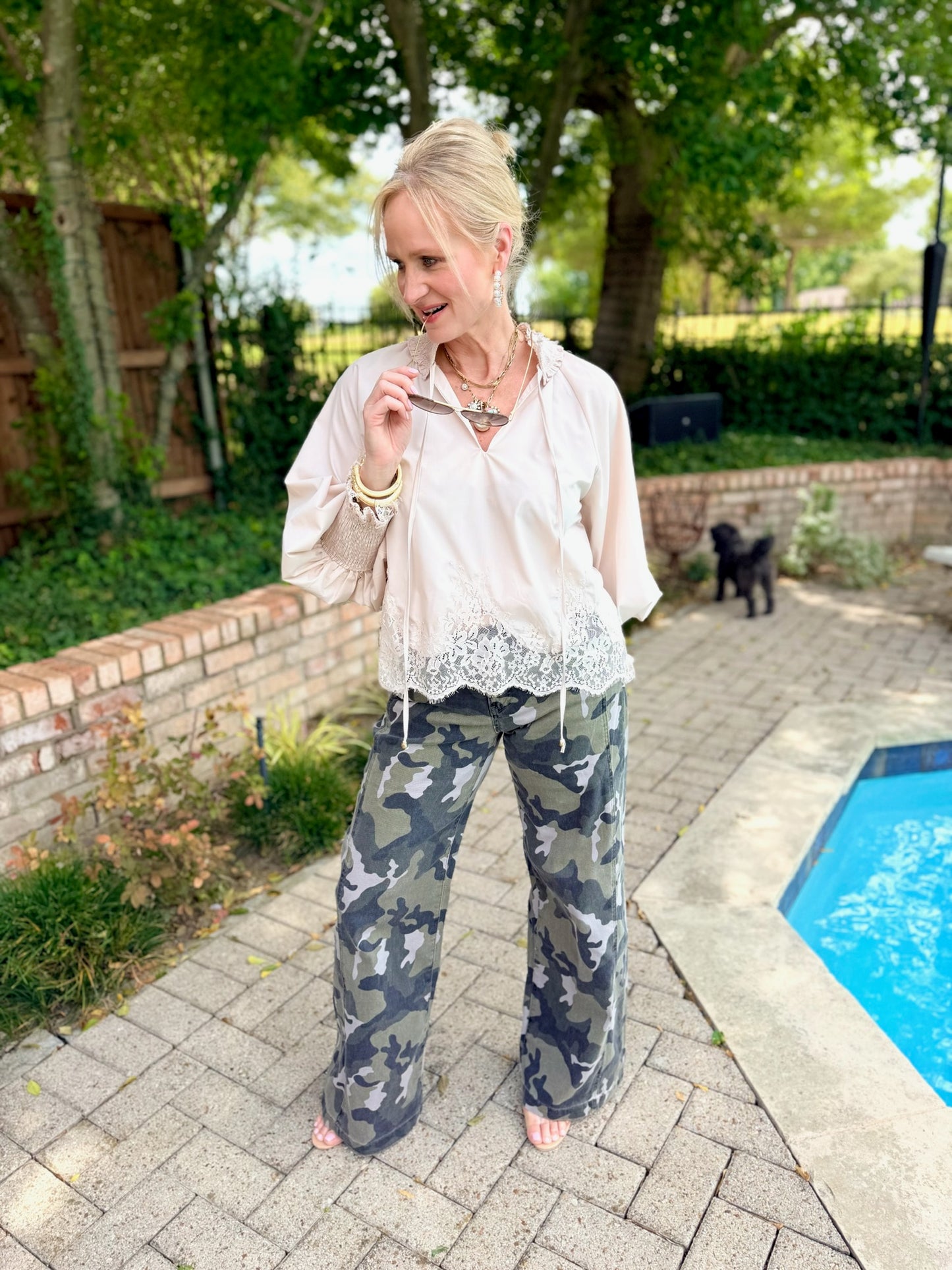 light champagne colored lace-trim blouse and camouflage wide-leg denim jeans . A small black dog is visible in the background near a brick wall and trees.