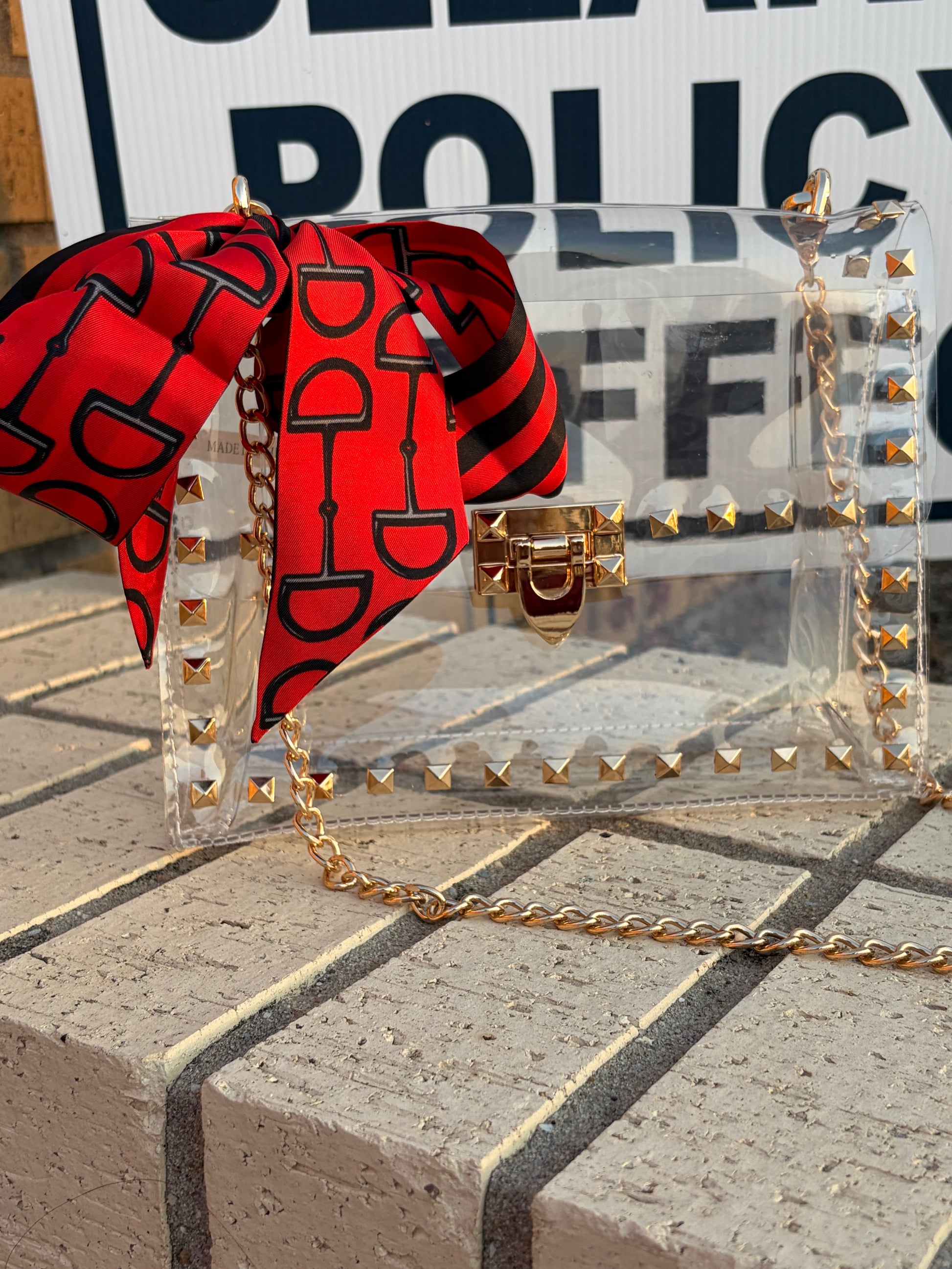 A clear game day shoulder bag with gold-tone studs and chain strap, styled with a red and black patterned silk scarf bow, resting on a stone surface.