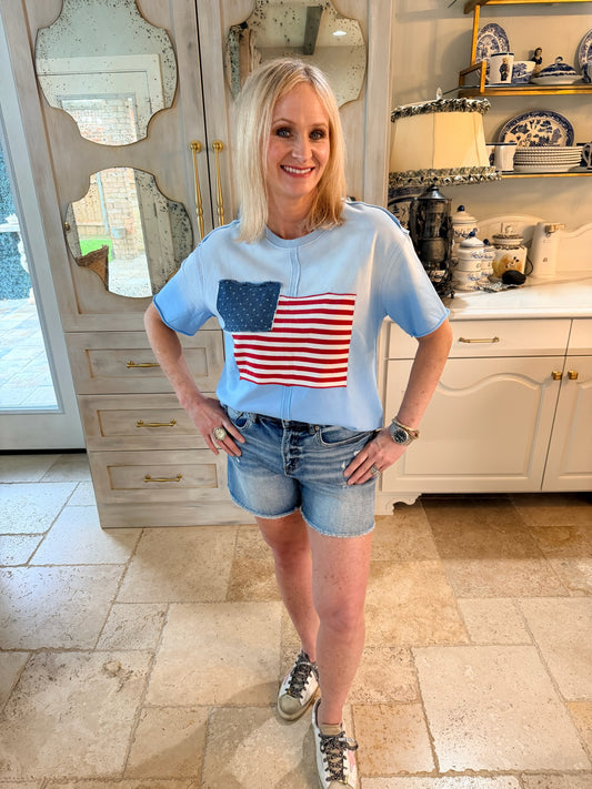 Woman wearing a light blue t-shirt with an American flag design in a kitchen.