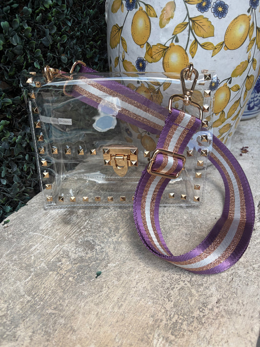 A clear plastic handbag with gold-tone studs and hardware, featuring a wide purple, white, and gold striped webbed wristlet strap, displayed outdoors on a stone surface.