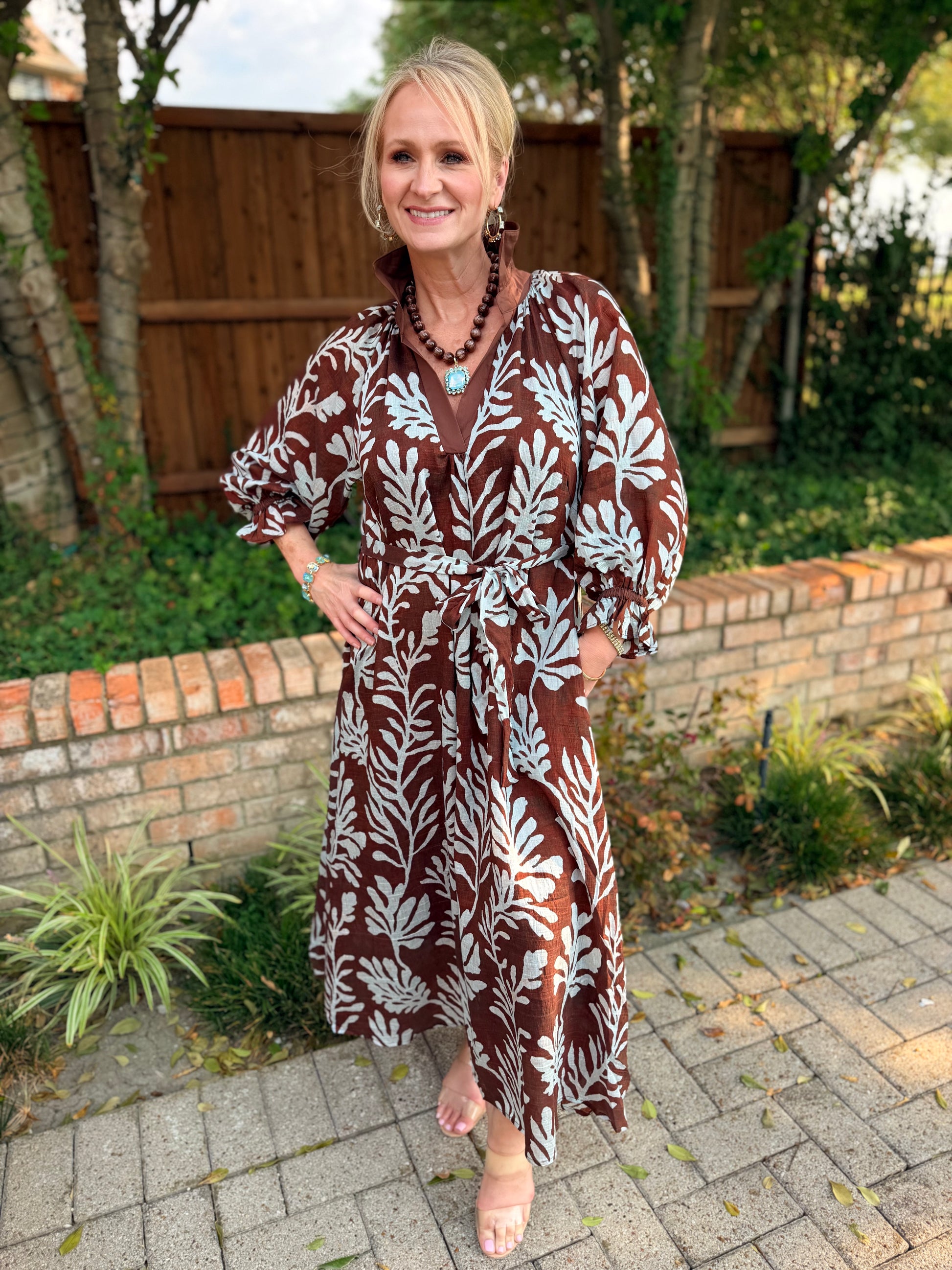 A woman wearing a brown and light blue botanical print midi dress, standing outdoors on a paved walkway with a brick wall and wooden fence in the background.