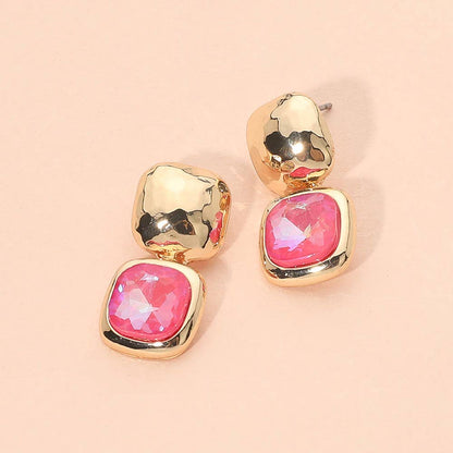 A pair of gold-toned drop earrings with a textured metallic top and a square pink faceted gemstone setting below, displayed on a solid pale pink background.