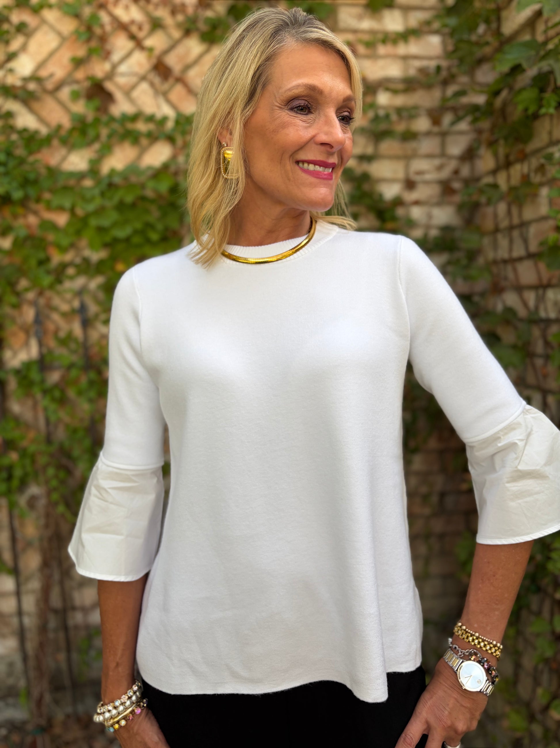 A woman wearing a white three-quarter sleeve top with ruffled cuffs, styled with a gold collar necklace and gold earrings, standing outdoors against a brick wall with green ivy.