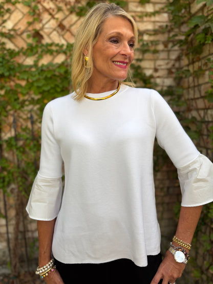 A woman wearing a white three-quarter sleeve top with ruffled cuffs, styled with a gold collar necklace and gold earrings, standing outdoors against a brick wall with green ivy.