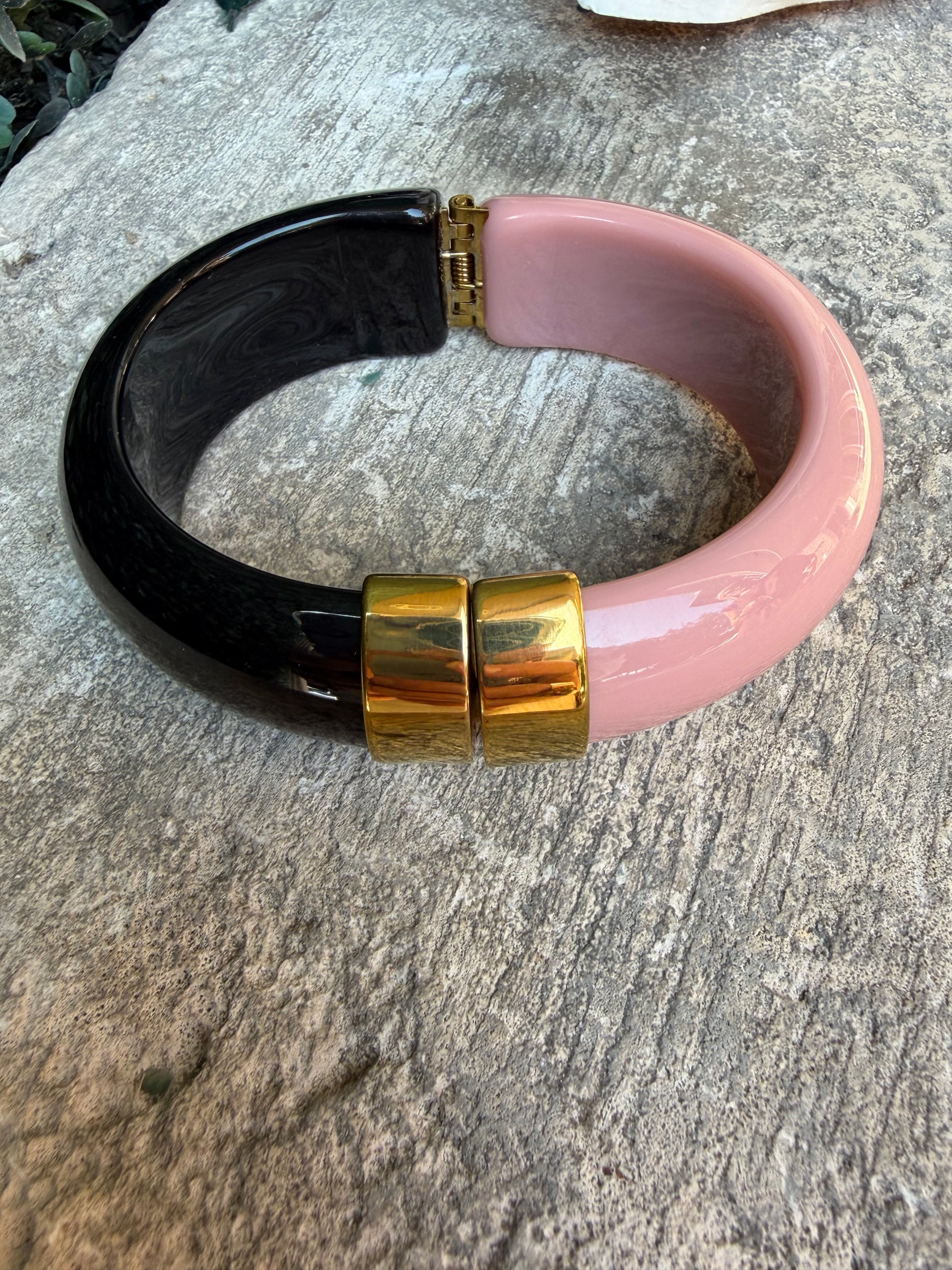 A chunky hinged bangle bracelet with a glossy pink upper section, a glossy black lower section, and two polished gold-tone metal hinge accents, displayed on a textured gray stone surface.