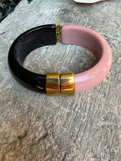 A chunky hinged bangle bracelet with a glossy pink upper section, a glossy black lower section, and two polished gold-tone metal hinge accents, displayed on a textured gray stone surface.