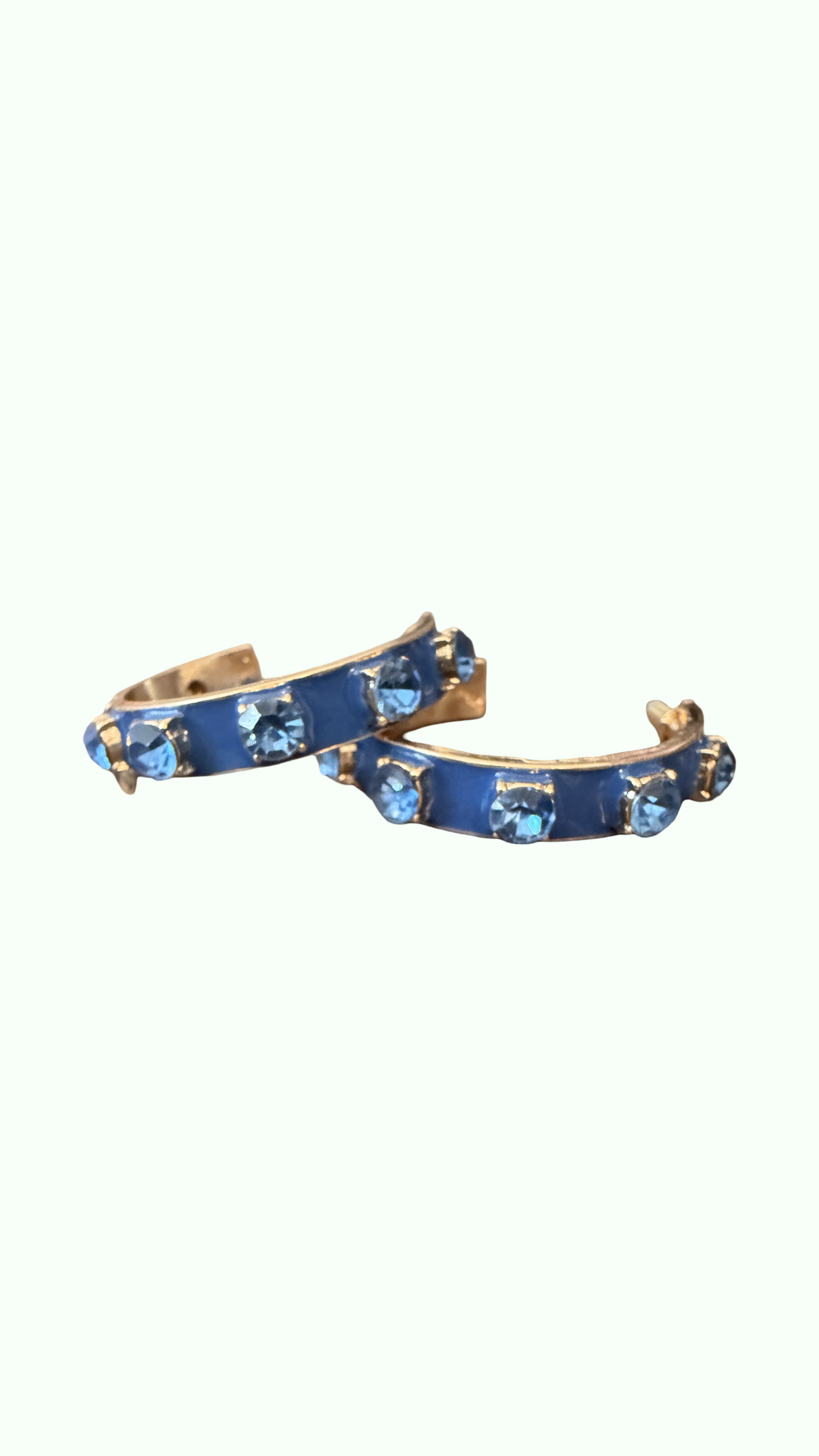 A pair of gold-toned hoop earrings with a vibrant blue enamel inlay and small light blue faceted gemstones, set against a solid off-white background.