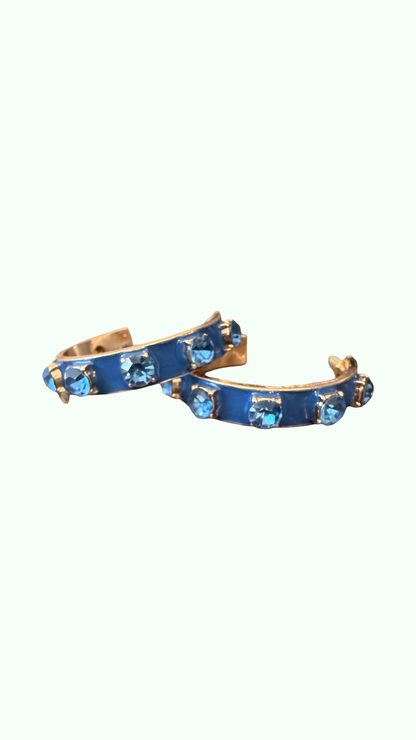 A pair of gold-toned hoop earrings with a vibrant blue enamel inlay and small light blue faceted gemstones, set against a solid off-white background.