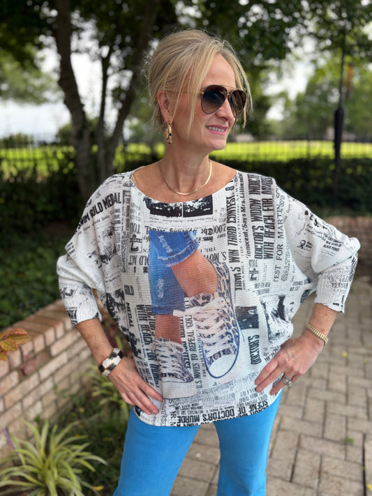 A woman wearing a white long-sleeve top with a black and gray newspaper print and a blue graphic patch, paired with bright blue pants, standing outdoors on a paved walkway with trees in the background.
