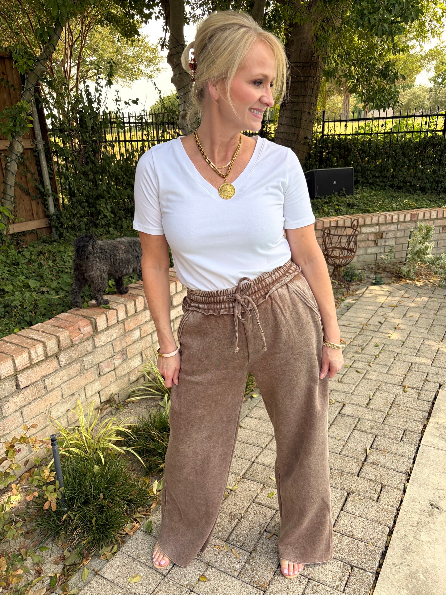 A woman standing outdoors on a paved walkway wearing a white short-sleeve V-neck t-shirt and mocha brown wide-leg drawstring cotton terry palazzo pant. 