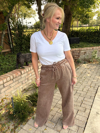 A woman standing outdoors on a paved walkway wearing a white short-sleeve V-neck t-shirt and mocha brown wide-leg drawstring cotton terry palazzo pant. 