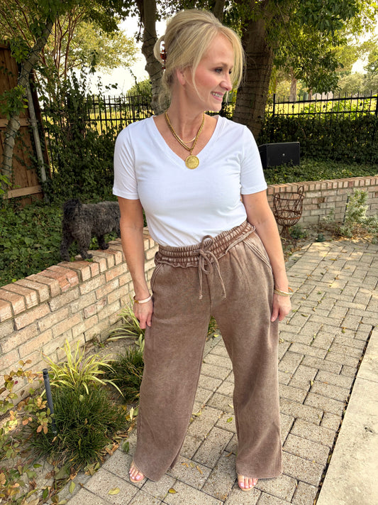 A woman standing outdoors on a paved walkway wearing a white short-sleeve V-neck t-shirt and mocha brown wide-leg drawstring cotton terry palazzo pant. 