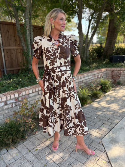 A woman wearing a brown and cream floral midi dress with puff sleeves and a tie waist, standing on a paved walkway in a backyard setting with trees and a brick wall.