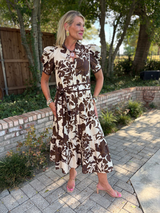 A woman wearing a brown and cream floral midi dress with puff sleeves and a tie waist, standing on a paved walkway in a backyard setting with trees and a brick wall.