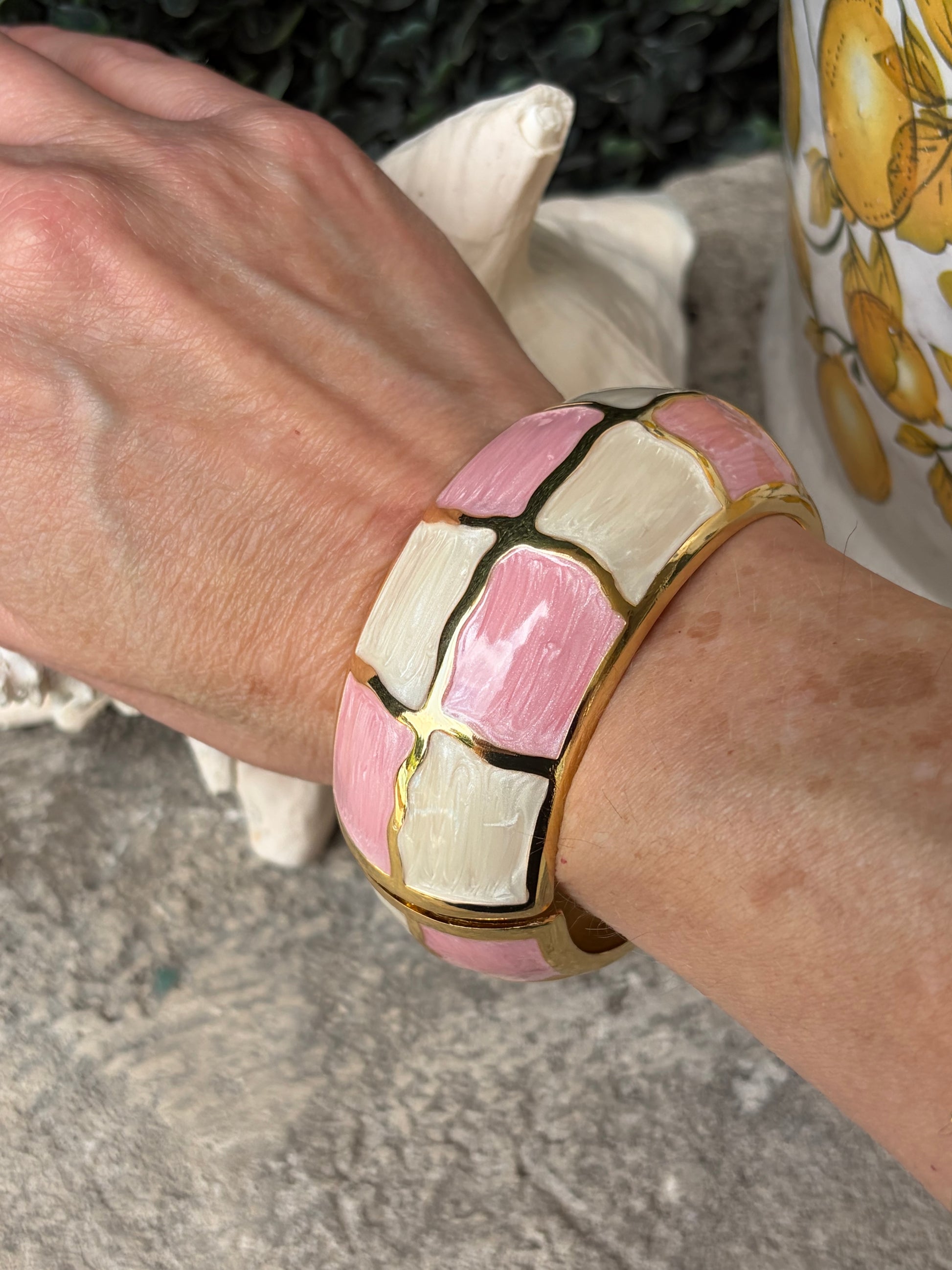 A close-up of a person's wrist wearing a chunky gold-tone bangle bracelet with a pink and cream mosaic tile pattern.