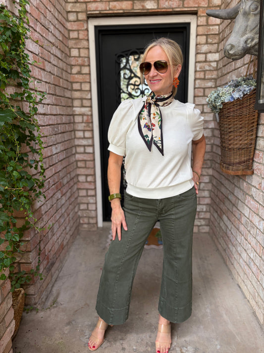 A woman stands in a brick walkway doorway wearing a cream puff-sleeve top with a floral neck scarf, olive green wide-leg pants, sunglasses, and red heels.