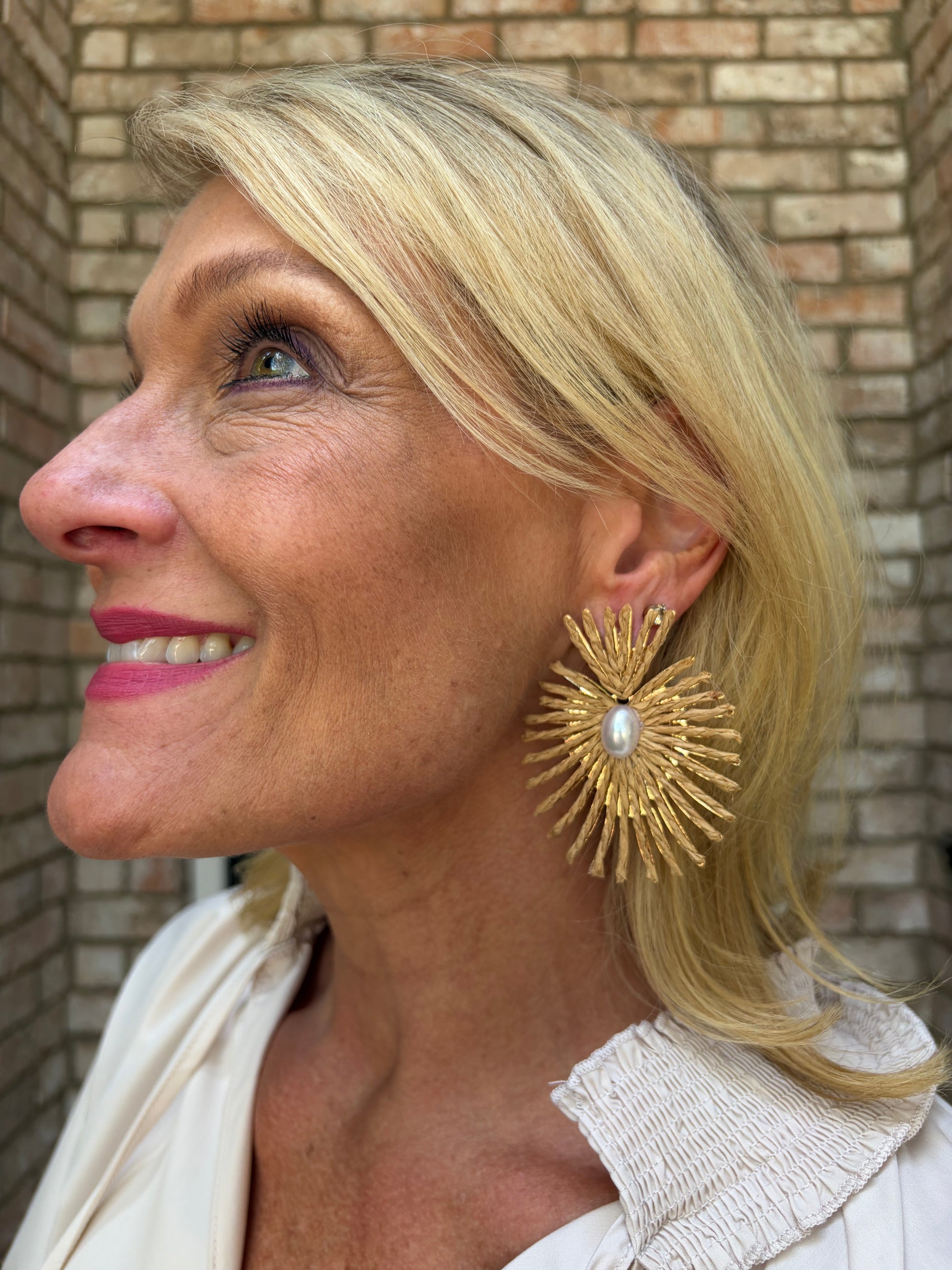 A close-up side profile of a smiling blonde woman wearing a large, ornate gold sunburst earring with a central white pearl, set against a brick wall background.