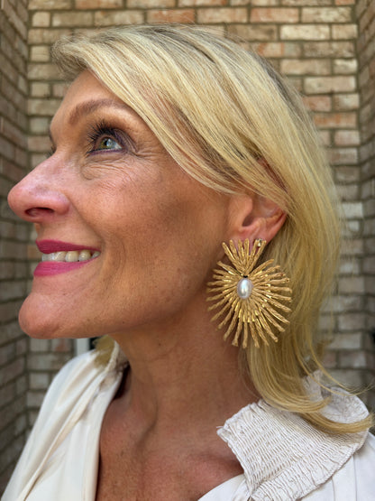 A close-up side profile of a smiling blonde woman wearing a large, ornate gold sunburst earring with a central white pearl, set against a brick wall background.