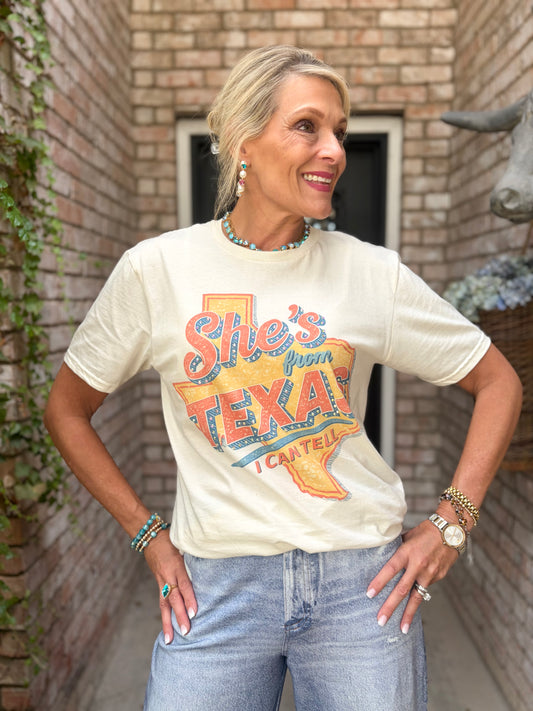 A woman wearing a cream-colored graphic t-shirt with a colorful 'She's from TEXAS” design, paired with light blue jeans, standing in front of a brick wall.