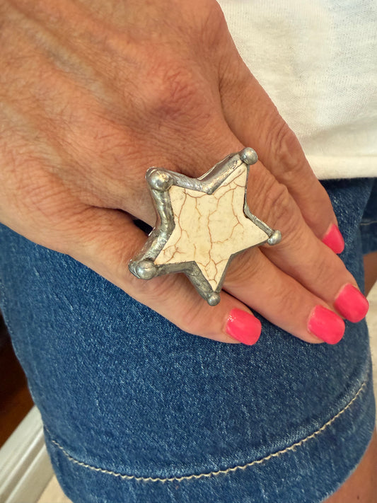 A close-up of a hand wearing a large silver-toned star-shaped ring with a textured cream inlay, styled with bright pink nail polish and blue denim shorts.