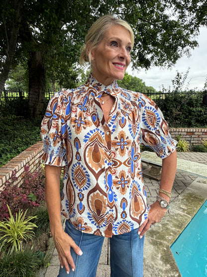 woman wearing brown and royal blue blouse with ruffled collar, button down, short sleeve and paired with light denim with a seam down the leg