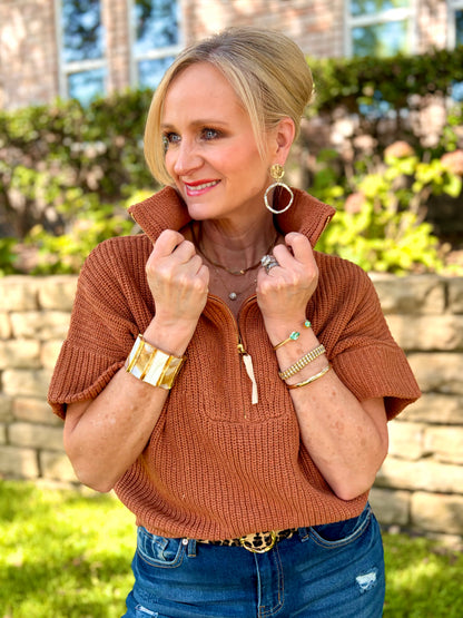 A woman wearing a pecan colored short-sleeve ribbed knit mock-neck sweater with a half-zip collar, styled with blue jeans and gold jewelry, standing outdoors in front of a stone wall and greenery.