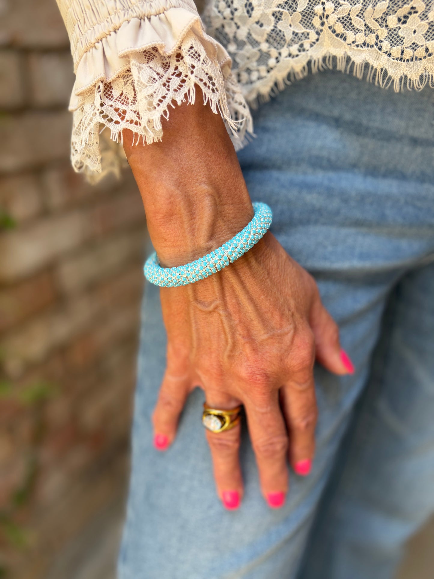 A close-up of a person's hand wearing a textured light blue beaded bangle bracelet, paired with a cream lace top and blue jeans.