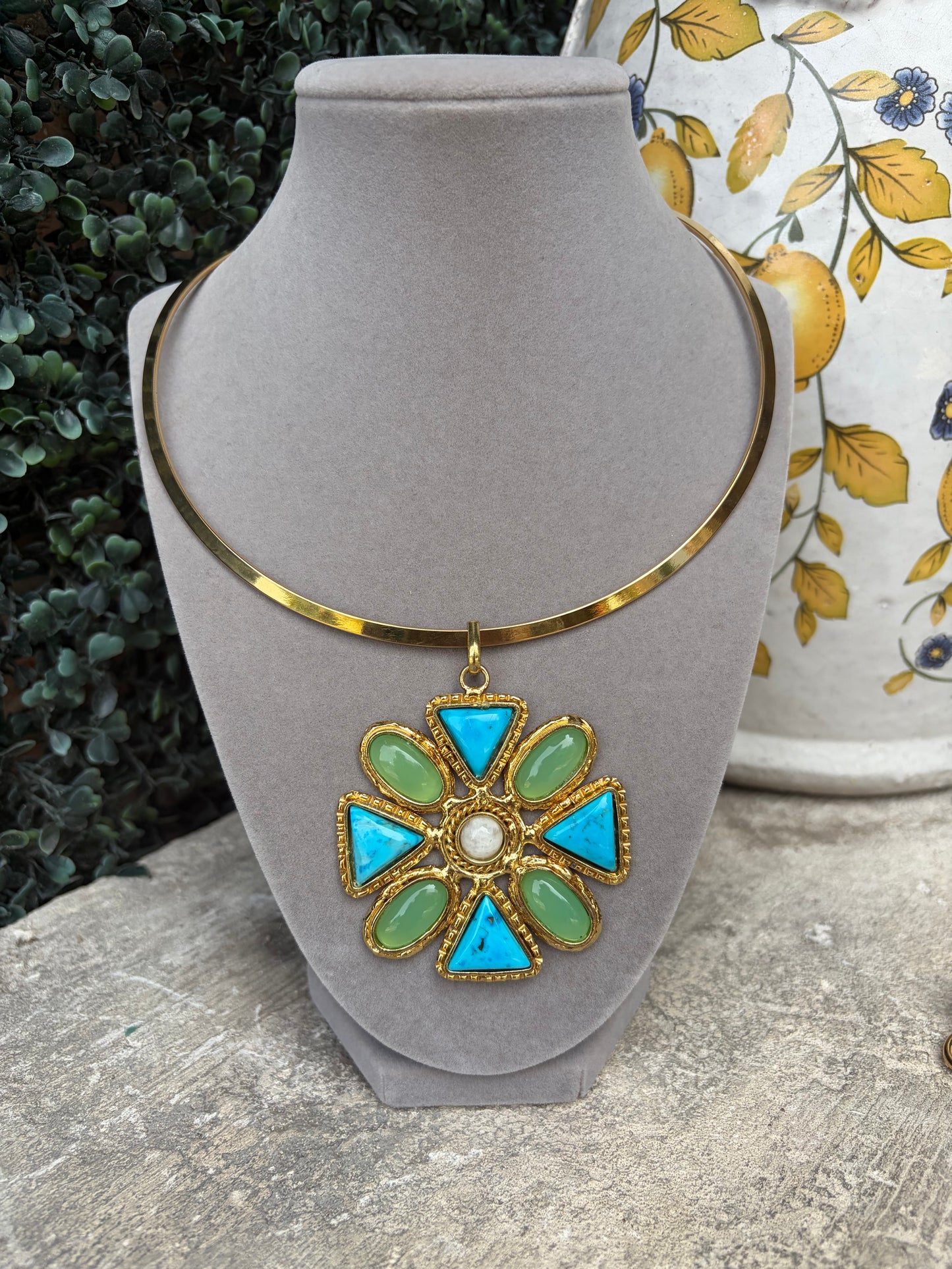 A gold-tone bangle necklace with a geometric pendant featuring turquoise blue and green stones, displayed on a gray velvet jewelry bust.