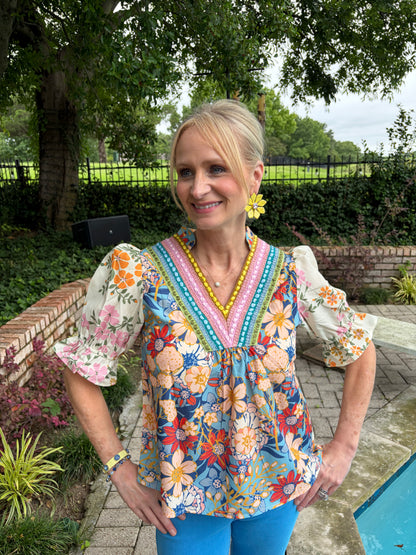 woman in a v neck that is embroidered, short sleeve blouse. floral with blues and yellows, orange and red. paired with a sea blue denim
