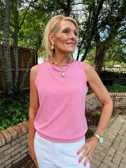 Woman wearing a pink sleeveless top and white pants standing outdoors with trees and a wooden fence in the background.