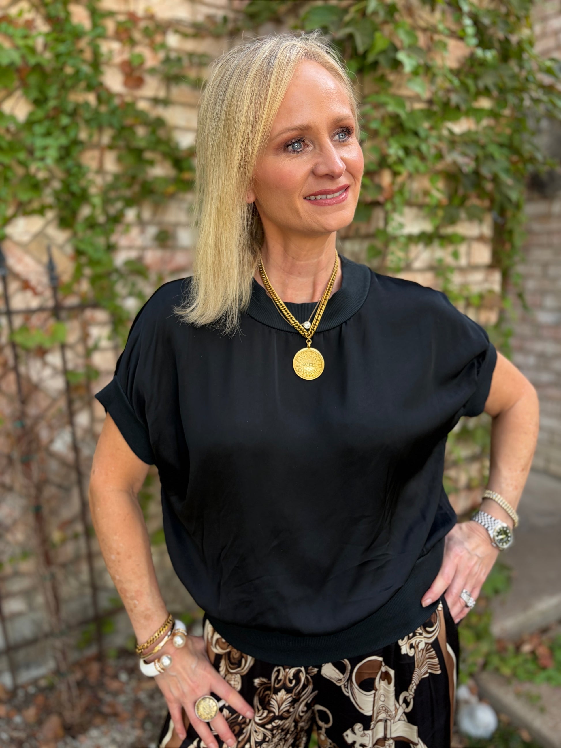 A blonde woman wearing a black short-sleeve top and a black-and-white patterned skirt, accessorized with gold necklaces and bracelets, standing outdoors in front of a brick wall with green ivy.