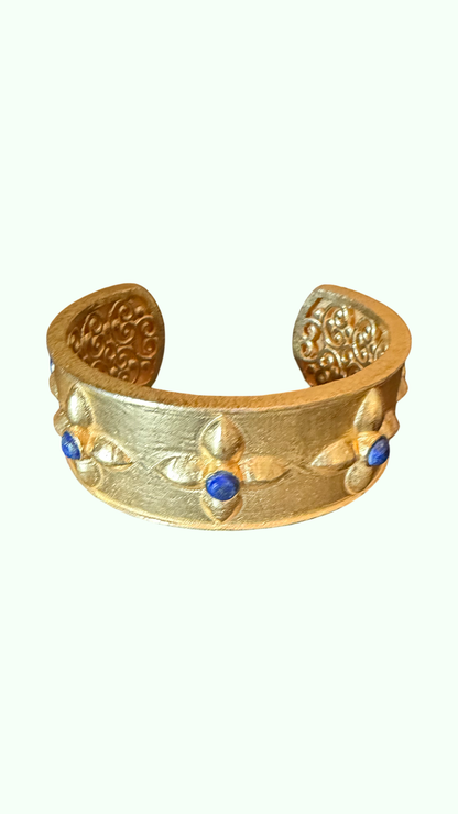A brushed gold-toned cuff bracelet with embossed clover and small blue stone accents in tarnish resistant