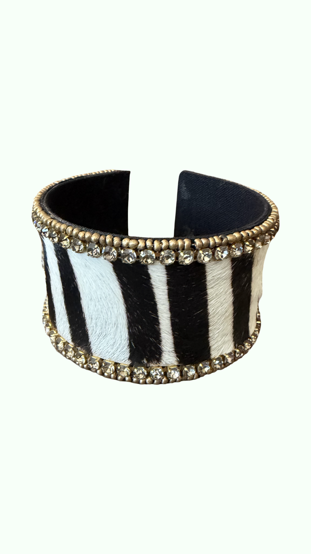 A wide cuff bracelet featuring a black and white zebra print hair-on-hide exterior, adorned with gold-tone beaded borders and clear rhinestones on both ends, set against a solid off-white background.